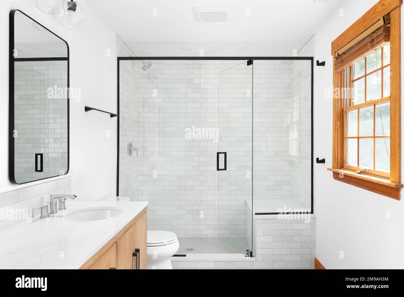 Une belle salle de bains moderne de luxe avec un cabinet en bois clair, une  douche à l'italienne avec des murs carrelés de métro en marbre, des  robinets et du matériel noirs