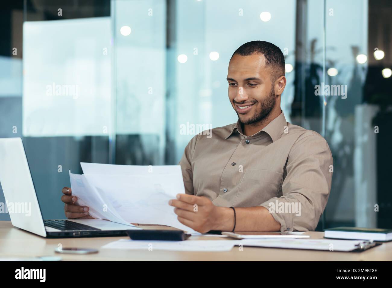 Homme d'affaires prospère dans le bureau lisant un rapport financier, homme hispanique tenant un contrat dans ses mains et souriant avec satisfaction, de bons résultats de réalisation, travaillant sur un ordinateur portable. Banque D'Images