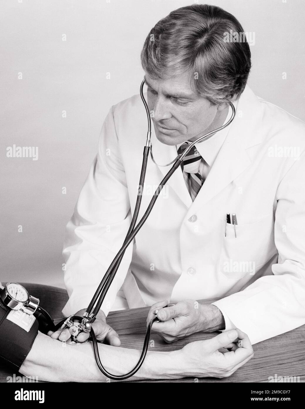 1970S MÉDECIN VÉRIFIANT LA TENSION ARTÉRIELLE D’UN PATIENT AVEC UN BRASSARD ET UN STÉTHOSCOPE - M9906 HAR001 PERSONNES HARS SOINS DU SANG HOMMES PROFESSION STÉTHOSCOPE CONFIANCE COMPÉTENCES EN SOINS DE SANTÉ B&W MANCHETTE COMPÉTENCES PROFESSIONNELLES BIEN-ÊTRE FOURNISSEUR DE PRÉVENTION ANGLE ÉLEVÉ MÉDECINS FOURNISSEURS PRATICIENS GUÉRISON ET DIAGNOSTIC CARRIÈRES MÉDECINS PROFESSIONNELS DE LA SANTÉ PROFESSIONNELS DE LA DÉFICIENCE TRAITEMENT GUÉRISSEUR DE LA PRESSION ARTÉRIELLE MÉDECIN CONCEPTUEL PRATICIEN SOUTIEN COOPÉRATION HOMME MOYEN-ADULTE PROFESSIONNELS NOIR ET LES MAINS DE LA MALADIE ETHNIQUE BLANCHE DE RACE BLANCHE NE SONT QUE HAR001 À L'ANCIENNE Banque D'Images