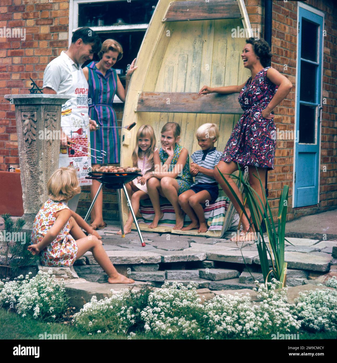 Une scène familiale heureuse comme Dad barbecue la nourriture sur un trépied barbecue sur le patio et trois des enfants utilisent un bateau à rames retourné comme siège! Banque D'Images