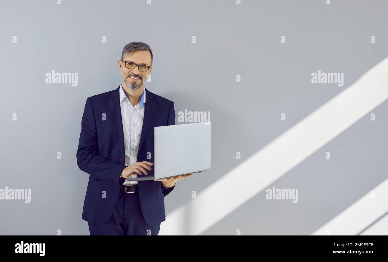 Portrait d'un homme d'affaires mûr heureux avec un ordinateur portable debout près d'un mur gris clair Banque D'Images