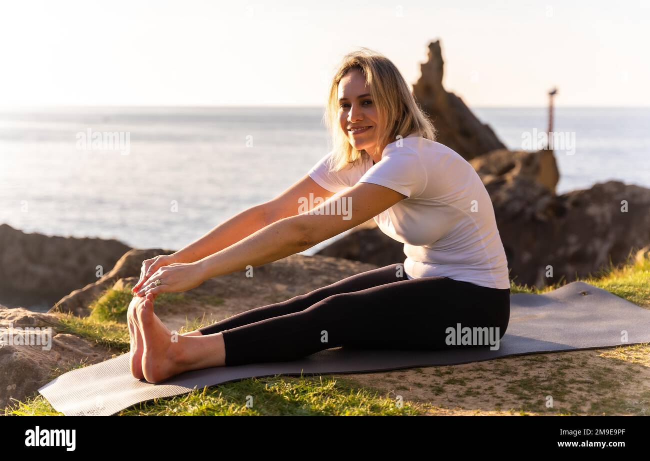 Une femme blonde faisant des exercices de yoga dans la nature au bord de la mer, s'étendant à côté d'un phare, la vie saine et naturiste, pilates à l'extérieur Banque D'Images