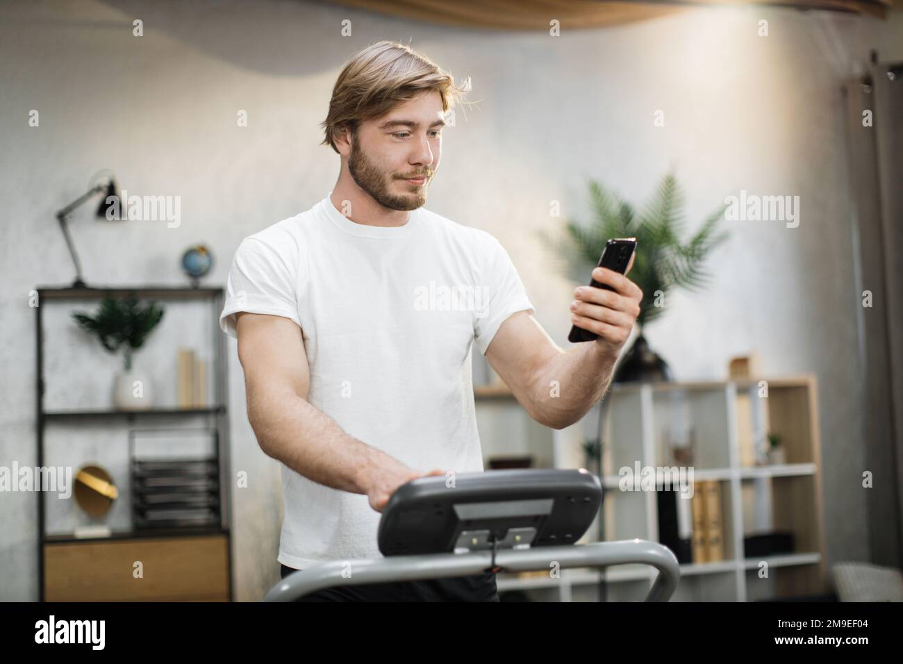 Gros plan sur un jeune sportif barbu à l'aide d'un simulateur de machine à  pied pendant un selfie ou un appel vidéo. Blonde sportif ayant  l'entraînement sur le tapis roulant à l'intérieur