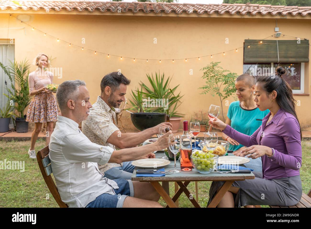 Multiculturel heureux amis riant avec un beau sourire autour de la table de déjeuner. Tandis que la femme apporte la nourriture à la maison patio. Banque D'Images