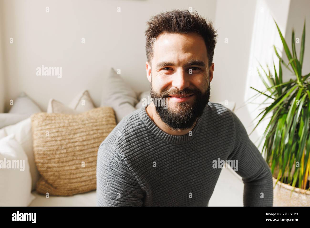 Portrait d'un jeune homme biracial à barbe souriante assis sur un canapé dans le salon au-dessus du mur, espace de copie Banque D'Images