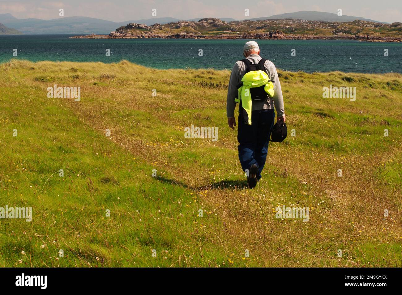 Man 60+ avec sac à dos et manteau de haute visibilité, marchant à travers l'herbe sur Iona vers la mer le son d'Iona, avec le Ross de Mull dans la distance Royaume-Uni Banque D'Images