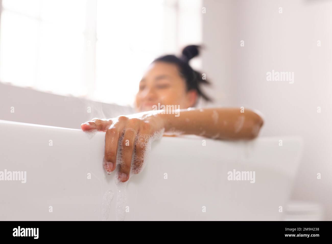 Image d'une femme biraciale souriante dans la baignoire dans le bain à bulles dans la salle de bains Banque D'Images