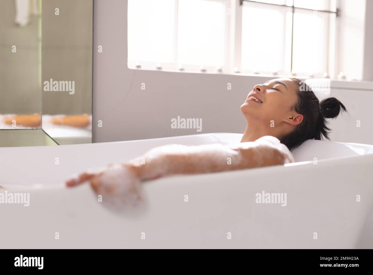Image d'une femme biraciale souriante dans la baignoire dans le bain à bulles dans la salle de bains Banque D'Images