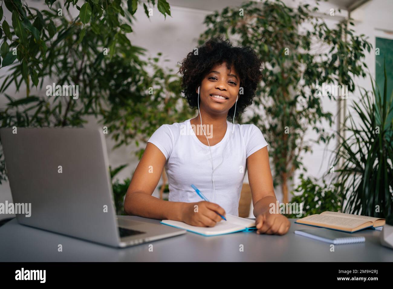 Portrait d'une étudiante afro-américaine joyeuse dans un casque tenant un stylo à la main assis au bureau avec un ordinateur portable, un livre en papier et souriant Banque D'Images