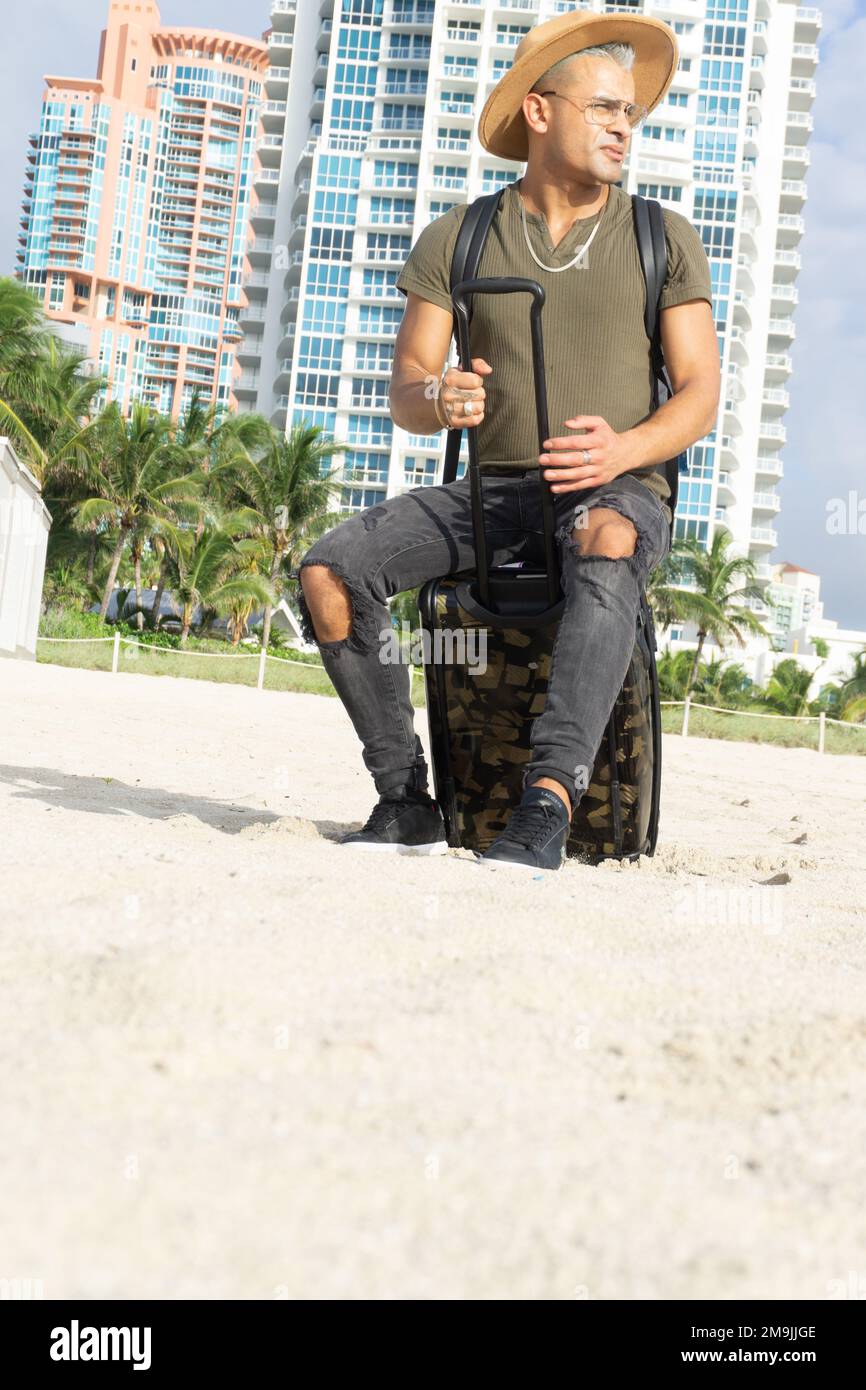 Gros plan d'un jeune homme faisant une pause sur son voyage. Un homme assis sur sa valise de transport sur la plage. South Beach, Miami. Concept voyage et été Banque D'Images