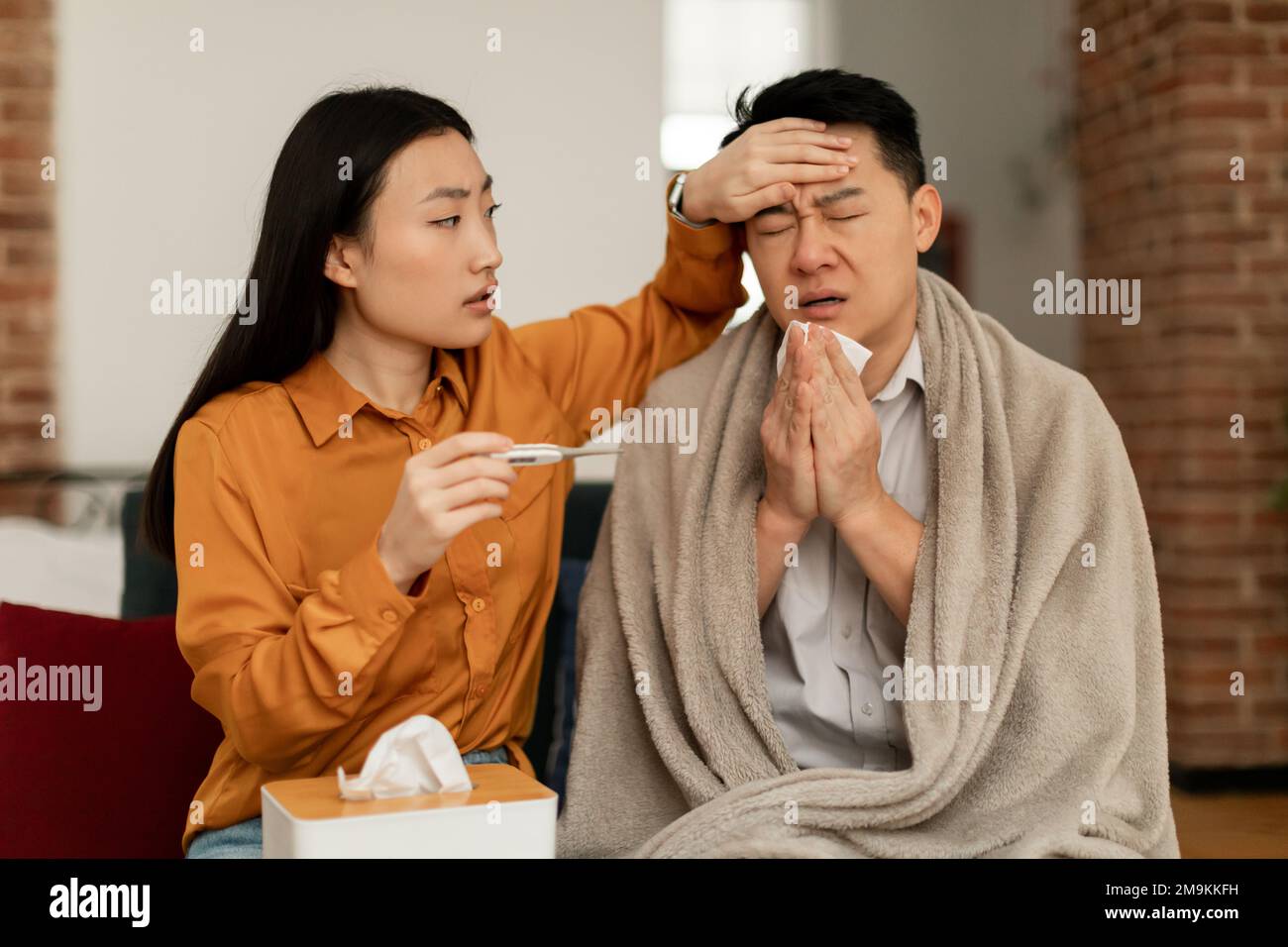 Femme asiatique traitant mari malade, tenant thermomètre et touchant le front de l'homme, assis sur un canapé à la maison Banque D'Images