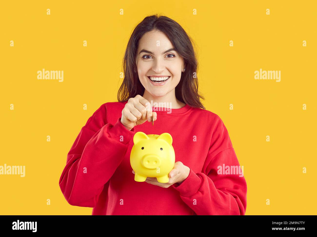 Portrait d'une femme brunette souriante et heureuse mettant de la pièce de monnaie dans une banque de porc jaune sur fond jaune. Banque D'Images