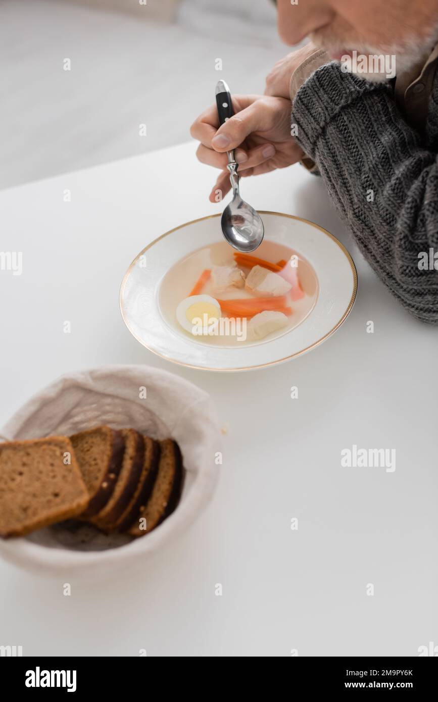 vue partielle de l'homme âgé souffrant de la maladie de parkinson et assis avec une cuillère près de la soupe et du pain dans la cuisine, image de stock Banque D'Images