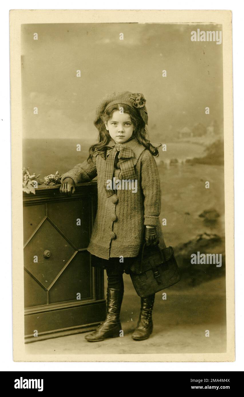 Magnifique original Edwardian Era Studio portrait carte postale de jeune fille édouardienne très mignonne avec de longs cheveux bouclés et portant un manteau tricoté et béret, tenant une mallette en cuir, écolière, circa 1910, Royaume-Uni Banque D'Images