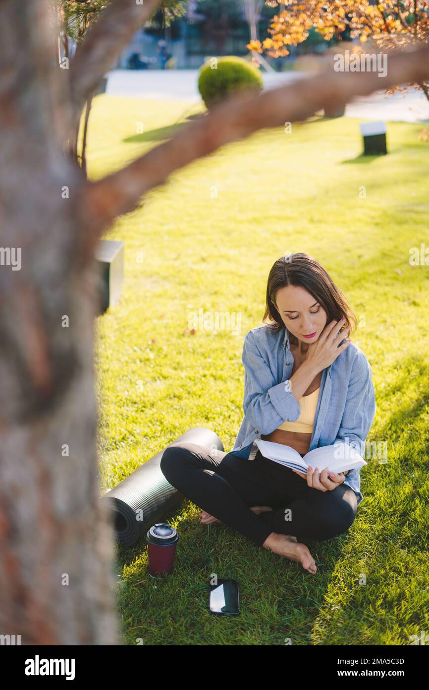 Jolie femme adulte portant des vêtements de sport lisant un livre avec enthousiasme, se reposant au parc après l'entraînement. Combinez le passe-temps de la lecture et du sport. Numérique Banque D'Images