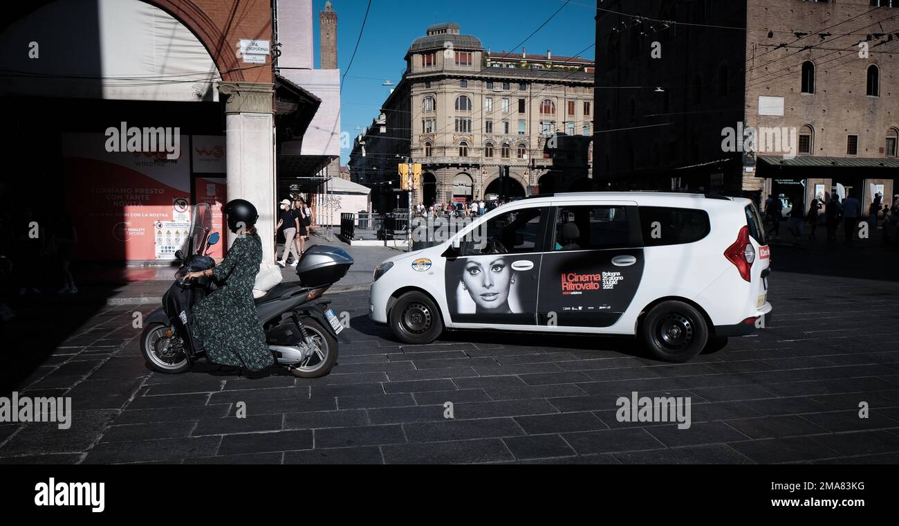 Une motard femme vêtue d'une robe se déplace à côté d'un taxi avec une publicité du cinéma Ritrovato avec une image de Sophie Loren à la porte de Bologne Banque D'Images