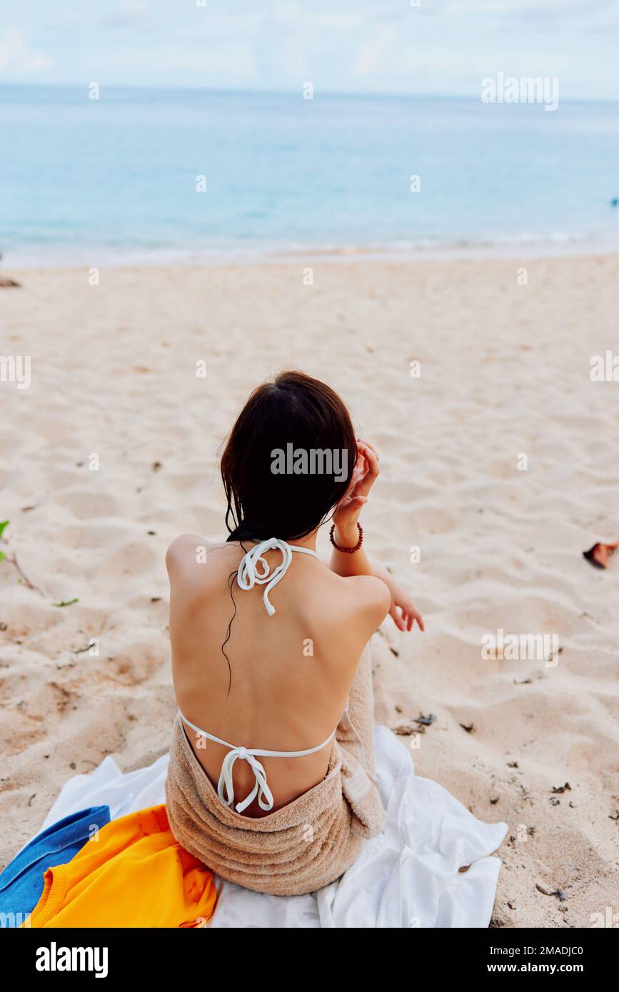 Une femme est assise avec elle dos à la caméra après avoir nagé dans l'océan dans un maillot de bain sur une serviette sur le sable et regarde la mer et les nuages, l'été Banque D'Images