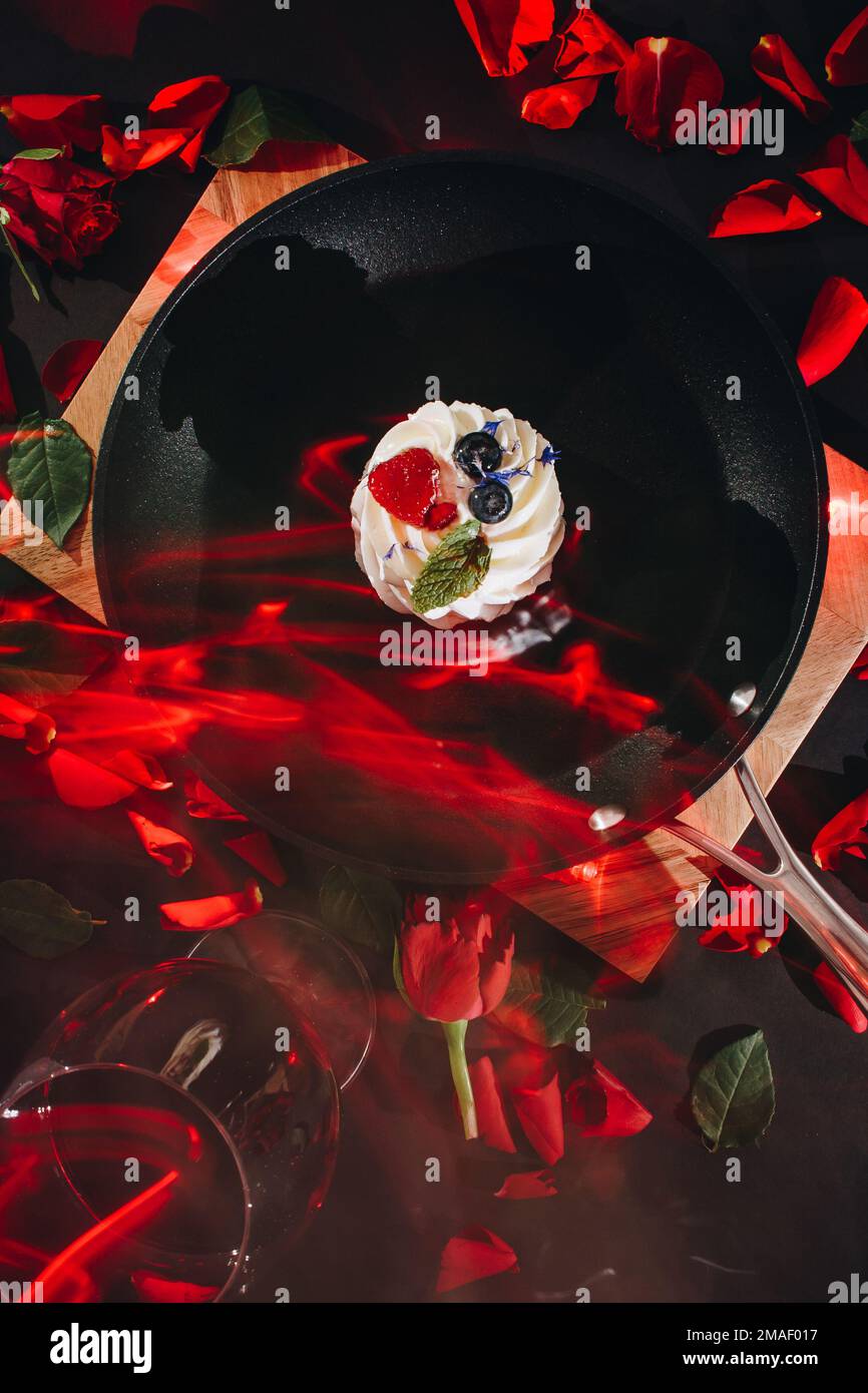 poêle à frire noire avec un gâteau pavlova et des pétales de rose rouges dispersés sur un panneau en bois avec un fond noir avec un verre de vin rouge et des bougies allumées Banque D'Images