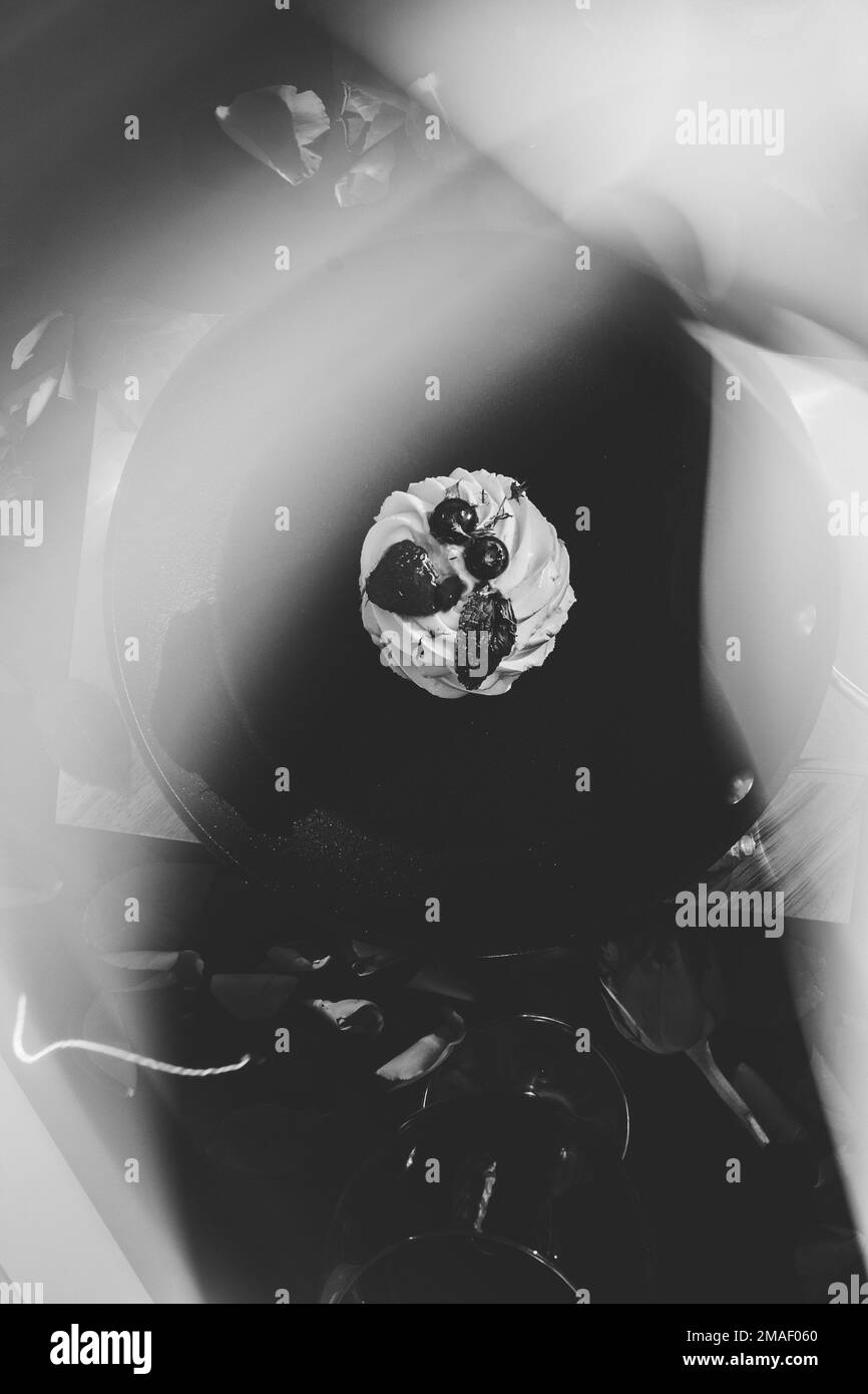 poêle à frire noire avec un gâteau pavlova et des pétales de rose rouges dispersés sur une planche de bois avec un fond noir à côté d'un verre de vin rouge et de fumée. fl Banque D'Images