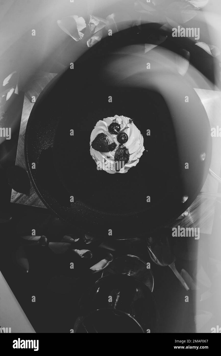 poêle à frire noire avec un gâteau pavlova et des pétales de rose rouges dispersés sur une planche de bois avec un fond noir à côté d'un verre de vin rouge et de fumée. fl Banque D'Images