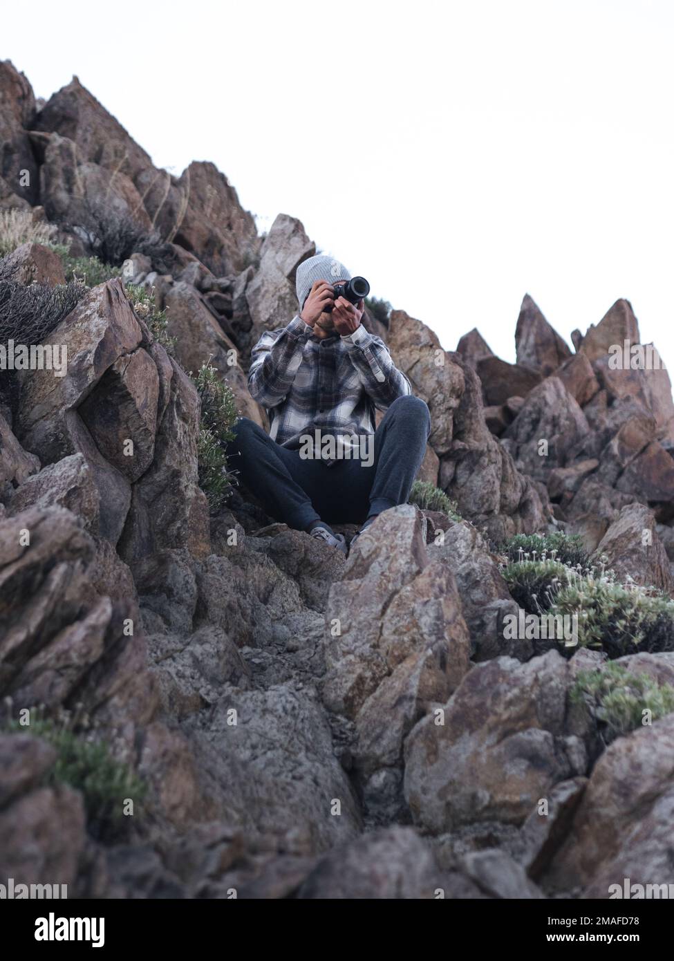 Jeune homme caucasien prenant des photos à l'ombre avec un appareil photo entouré de rochers dans une veste en flanelle et un bonnet. Banque D'Images
