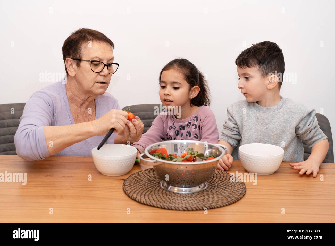 Une femme âgée et deux petits enfants dans la cuisine coupant la salade de fruits.grand-mère et petits-enfants cuisent la confiture de fraises ensemble.grand-mère enseignant aux enfants couper le dessus des fraises. Banque D'Images