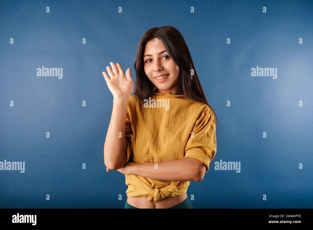 Femme brune portant un t-shirt jaune moutarde isolée sur fond bleu en souriant largement tout en agitant la paume relevée, saluant l'ami. Dire l'enfer Banque D'Images