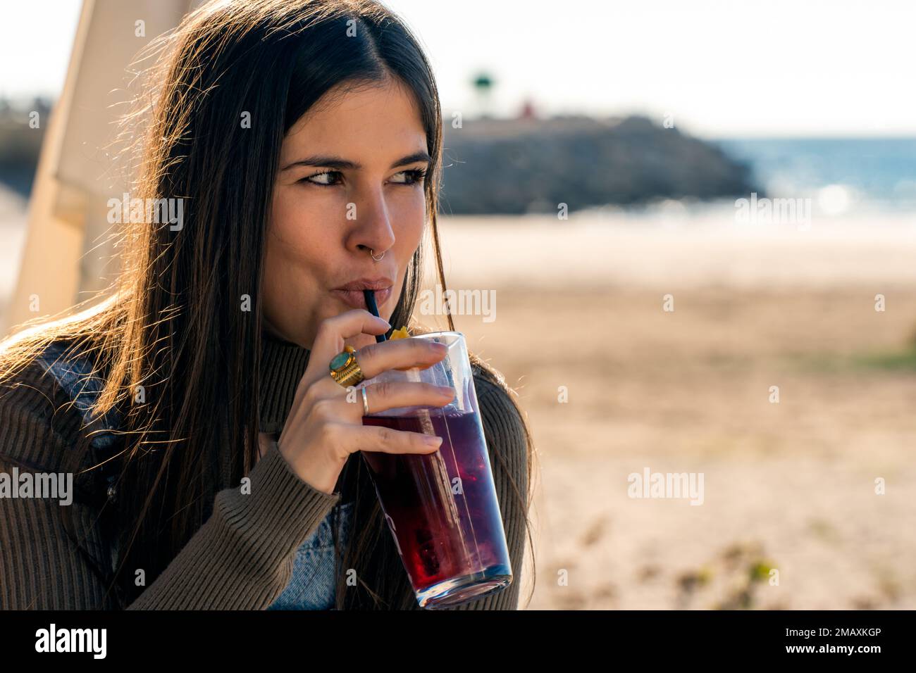 Jeune femme en vêtements décontractés buvant de la boisson fraîche rouge froide à travers la paille du verre tout en passant du temps sur la mer par temps ensoleillé Banque D'Images