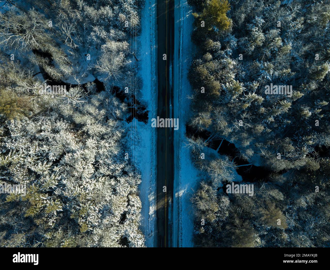 Une photo aérienne élevée centrée au-dessus d'une route droite à deux voies qui passe au-dessus d'un ruisseau dans une zone rurale et qui traverse une forêt enneigée et gelée. Banque D'Images