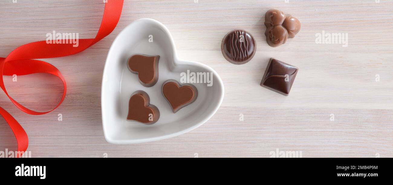 Arrière-plan avec assortiment de bonbons au chocolat dans un récipient en céramique blanche en forme de coeur sur une table en bois et un noeud rouge. Vue de dessus. Banque D'Images