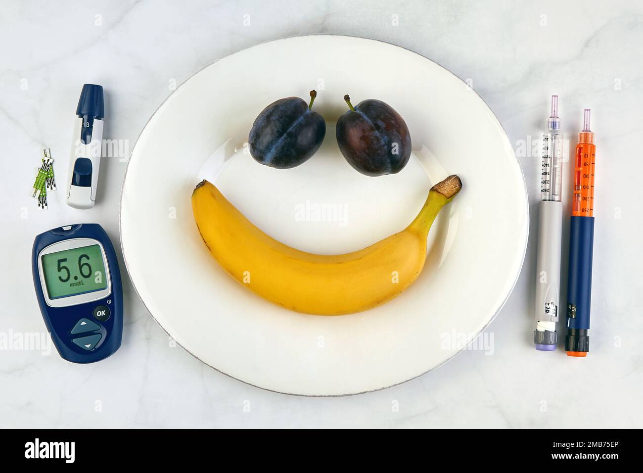Banane et prunes sous forme de smiley visage sur une plaque blanche et des stylos à seringue d'insuline et un glucomètre comme coutellerie sur une table en marbre Banque D'Images