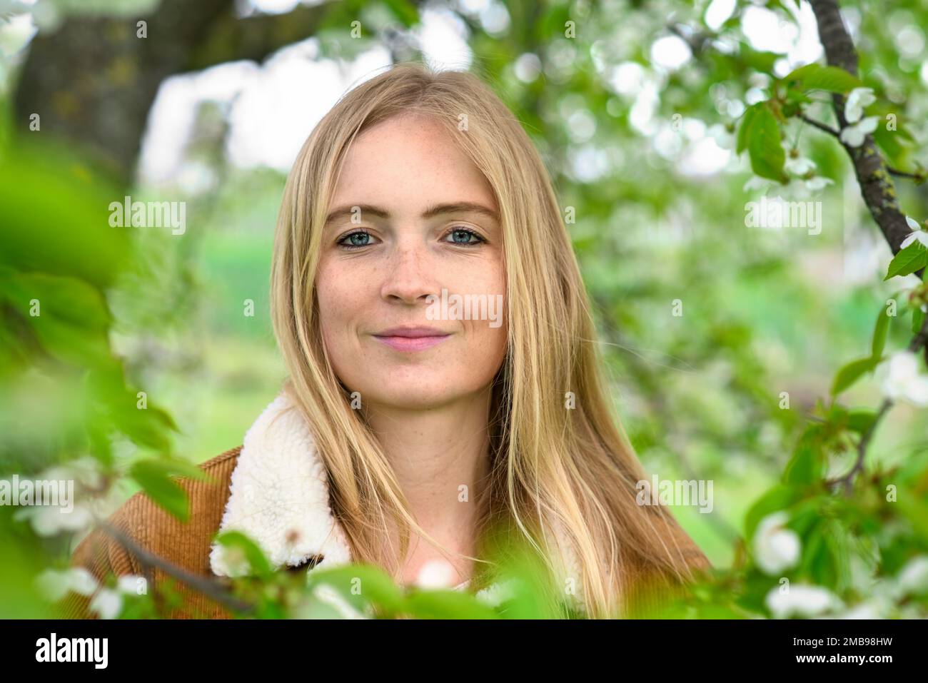 Portrait d'une belle jeune femme souriante dans la nature, avec des fleurs printanières et un feuillage vert tendre sur les branches d'arbre entourant son visage Banque D'Images