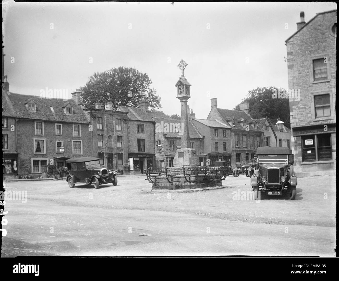 Place du marché, Stow-on-the-Wold, Cotswold, Gloucestershire, 1928. Une vue du sud-ouest à l'extrémité sud de la place du marché, montrant deux voitures garées à côté de la croix du marché avec des maisons et des magasins au-delà. Banque D'Images