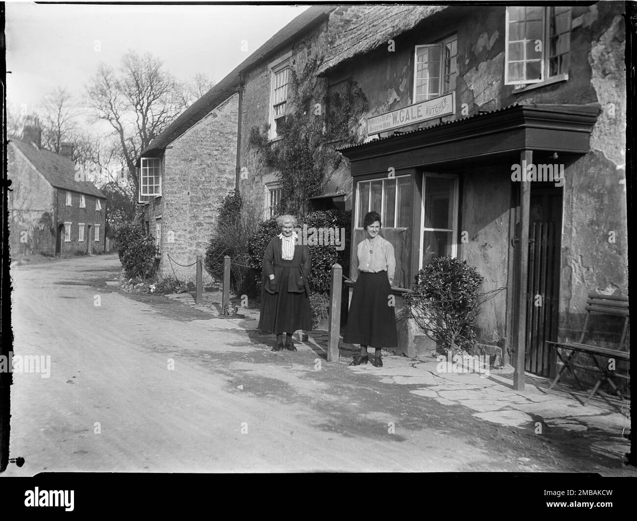 Hydrangea House, Grove Road, Burton Bradstock, West Dorset, Dorset, 1922. En regardant vers l'est sur Grove Road le long de l'avant de la boutique du boucher, montrant Mme Gale et Ivy debout à l'extérieur. Dans l'indice négatif de la collection, le photographe a enregistré l'emplacement de la photo sous le nom de Hydrangea House, qui semble être sur le site du 36-37 Grove Road et qui comprenait un boucherie. La photographe séjournait ici lors de sa visite à Burton Bradstock avec Mme Gale, qui était la femme du boucher. Banque D'Images