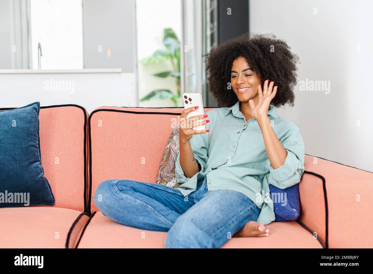 Une femme joyeuse et excitée, souriante, utilise un smartphone pour la connexion vidéo. Femme noire en tenant le téléphone mobile et en agitant, parlant sur le loin canapé assis à la maison Banque D'Images