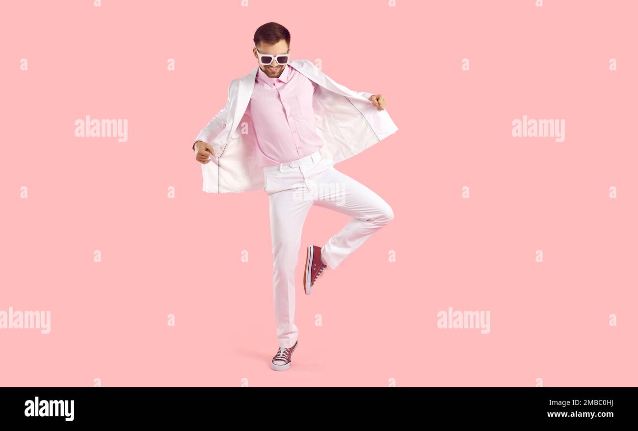 Drôle de gars en costume blanc, lunettes de soleil et entraîneurs dansant sur fond rose studio Banque D'Images