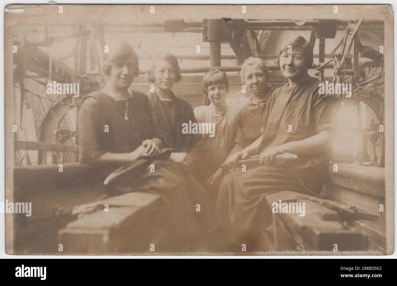 Photo du début du 20th siècle de cinq femmes ouvriers d'usine montré assis sur des bancs dans l'usine entre les rangées de métiers à coton. Trois générations de femmes sont incluses dans la photo. Deux des femmes tiennent des navettes de tissage de métier à tisser Banque D'Images