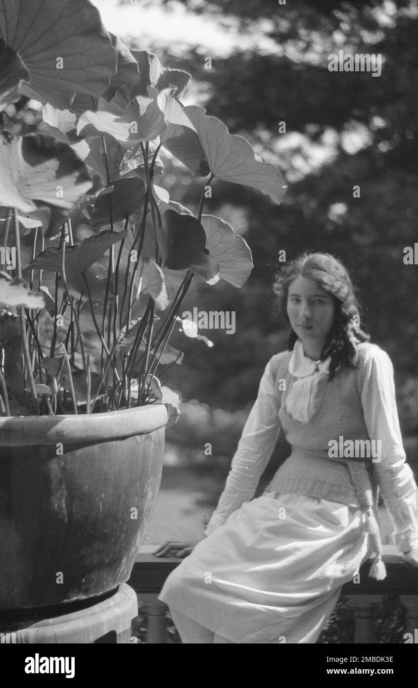 Femme non identifiée assise dans un jardin à côté d'une grande plante en pot, entre 1917 et 1934. Banque D'Images