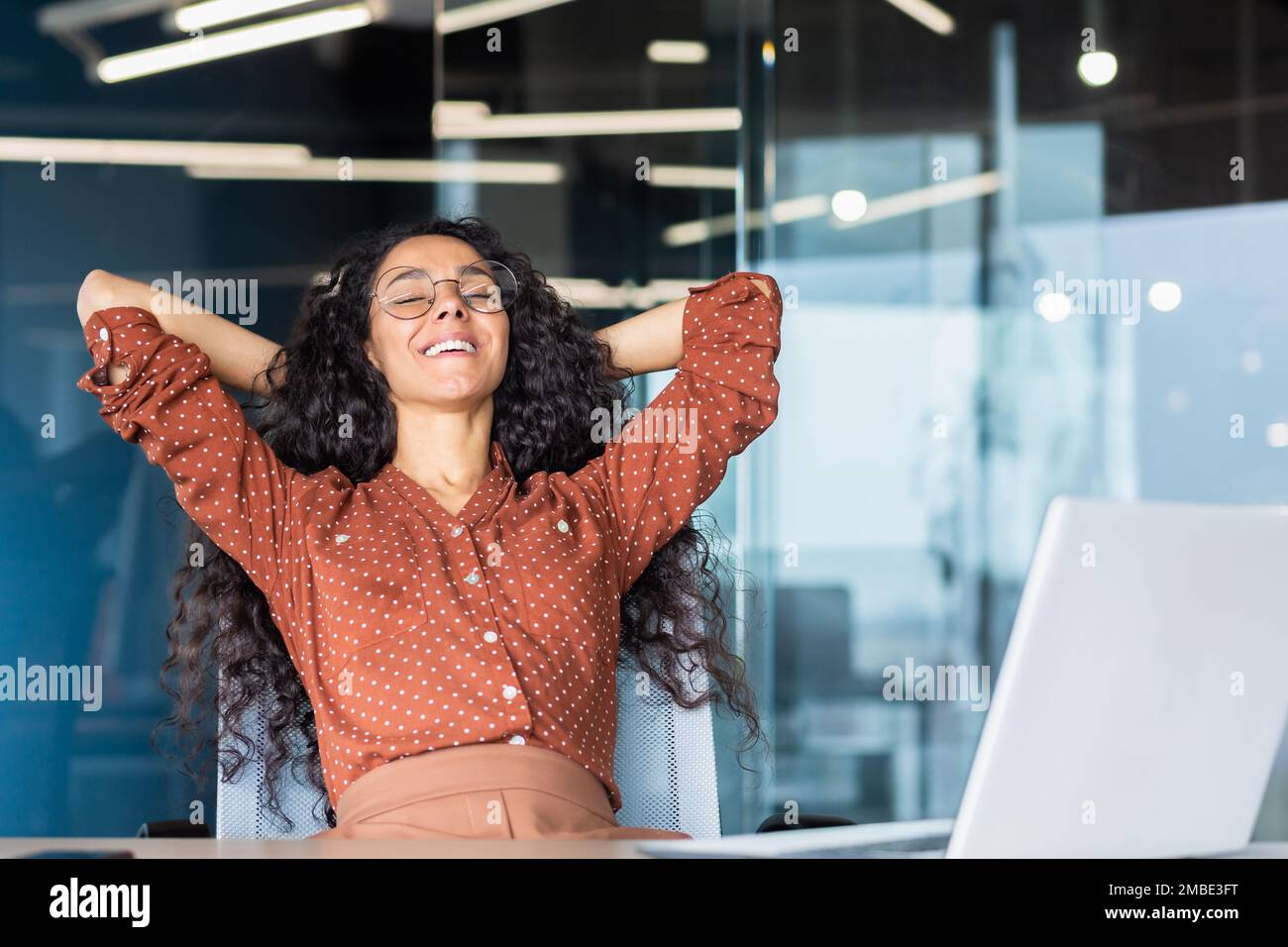 Une jeune belle étudiante latino-américaine est assise au bureau, sur le campus à une table avec un ordinateur portable, met ses mains derrière sa tête, ferme les yeux, se repose d'étudier, sourit. Banque D'Images