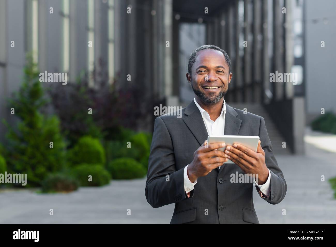 Portrait d'un patron afro-américain réussi, un homme d'affaires en costume souriant et regardant la caméra, un homme tenant une tablette informatique dans ses mains, un investisseur réussi naviguant sur Internet. Banque D'Images