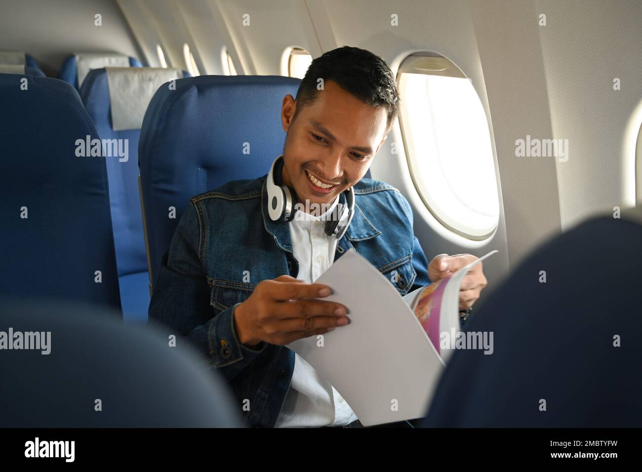 Homme asiatique gai passager dans des ensembles de hipster assis près de la fenêtre dans un avion et de lecture magazine Banque D'Images