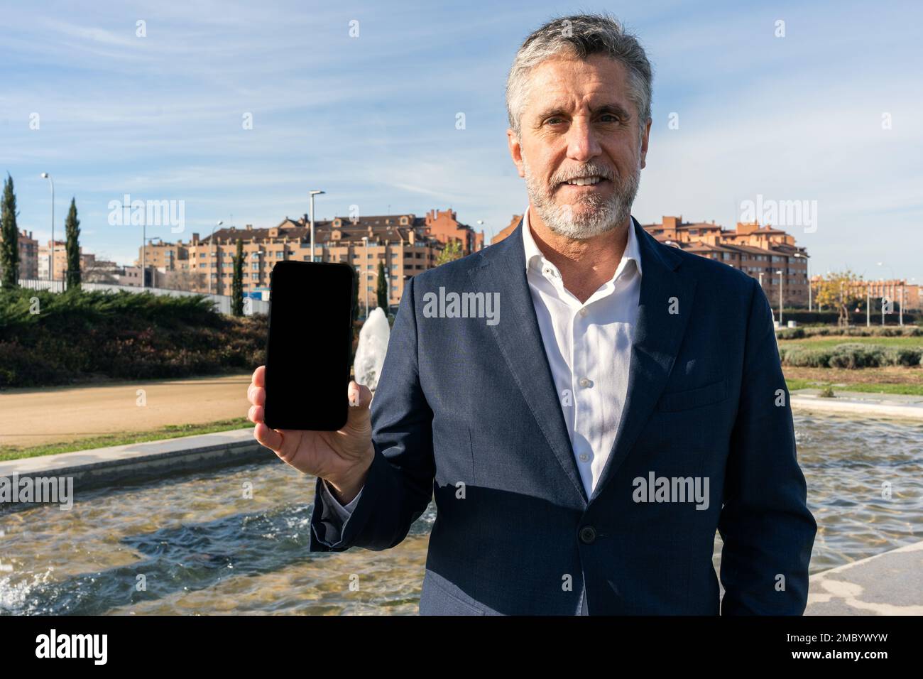 Homme d'affaires d'âge moyen positif dans une tenue habillée souriant et regardant l'appareil photo tout en montrant un smartphone avec écran noir dans la rue Banque D'Images