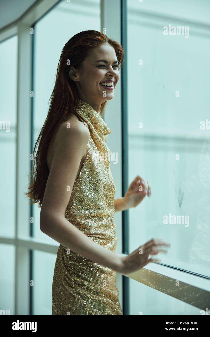 Belle femme de mode riant et souriant dans une élégante chère robe à paillettes dorées pose donnant sur la fenêtre de la ville dans un hôtel, Noël Banque D'Images