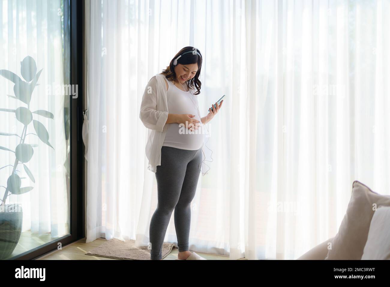 Portrait d'une femme asiatique heureuse enceinte debout dans le salon à la maison et utilisant un smartphone pour écouter de la musique dans un casque. Banque D'Images