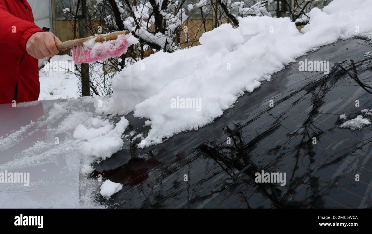 un homme dans une veste rouge nettoie le pare-brise d'une voiture avec un reflet de branches sur le verre d'une couche de neige à l'aide d'une brosse maison Banque D'Images