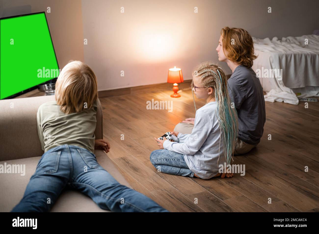 Trois enfants jouant au jeu vidéo Green Screen chromakey sur la console de télévision à l'aide de la manette tout en étant assis à la maison, frères et soeur assis sur le sol Banque D'Images