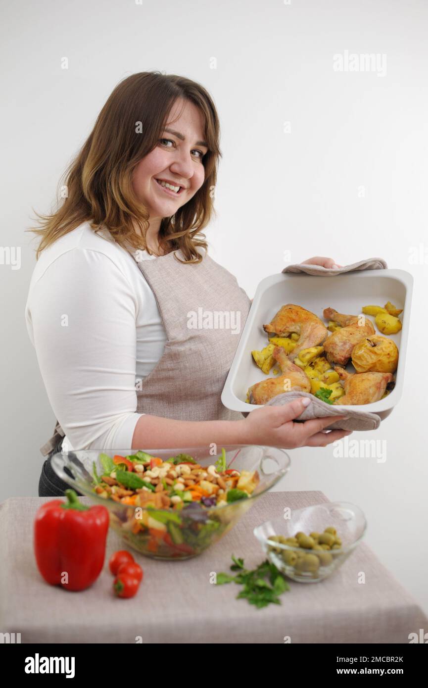 adulte belle plump femme en tablier prépare le dîner sur la plaque de cuisson des jambes de poulet quarts de pommes de terre à proximité grand bol en verre avec la salade et les noix différents ingrédients elle sourit Banque D'Images