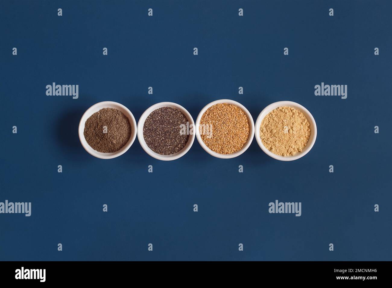 Quatre bols en céramique blanche dans une rangée avec des graines de chia et de lin entières et en poudre sur un fond bleu studio et un espace de copie. Santé et superalimentation. Banque D'Images