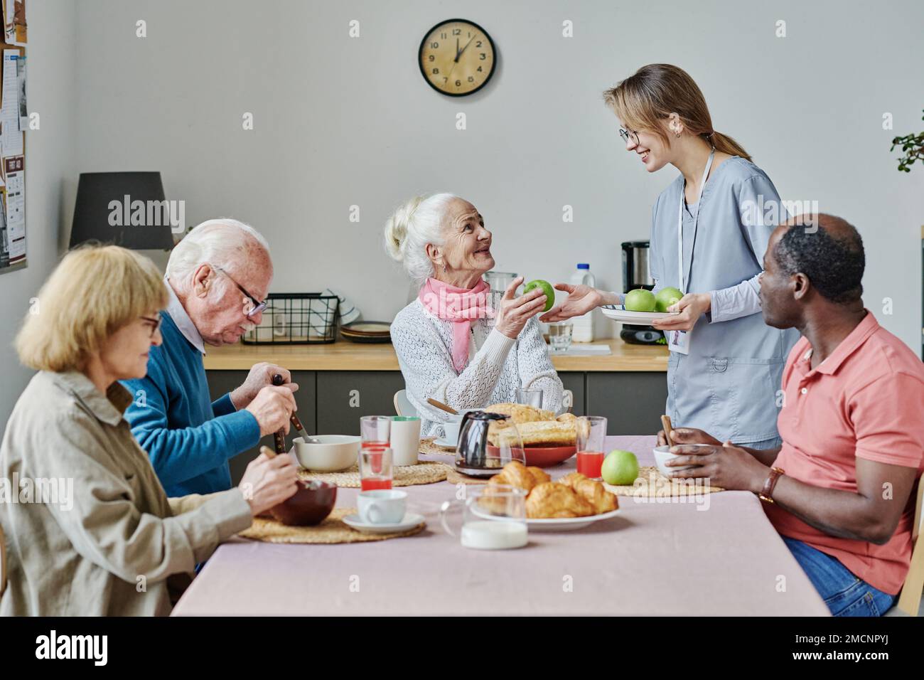 Les jeunes souriants se portent volontaires pour traiter les pommes aux personnes âgées pendant qu'elles mangent à table dans la cuisine du foyer de soins infirmiers Banque D'Images