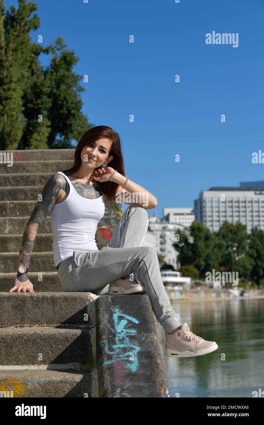Jolie jeune femme confiante avec des tatouages frappants se trouve sur un escalier près d'une rivière dans la ville Banque D'Images